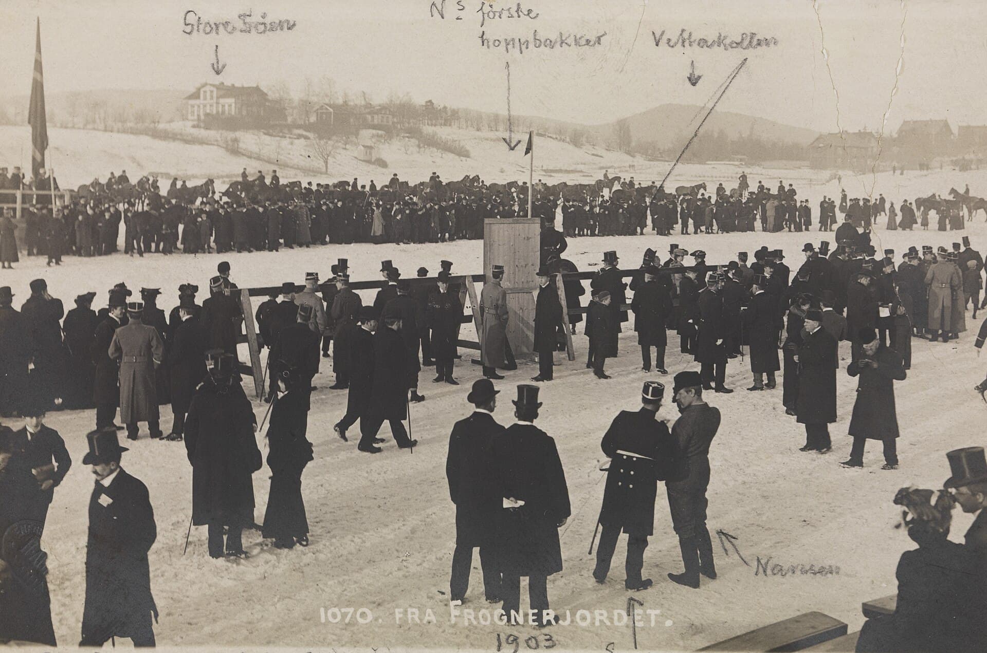 Crowd and competitors gathered on a snowy field while two men speak in the foreground.